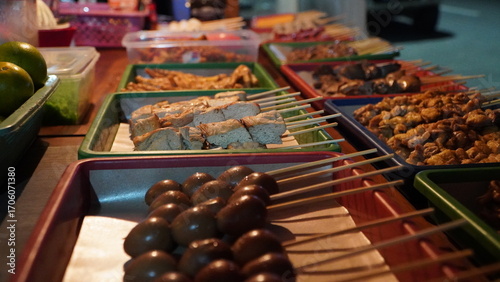 street food stall with skewered items including meat, tofu, and eggs neatly arranged in trays on table, outdoor market setting with background containers and vehicle