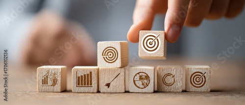 Hand placing a wood block with a target icon on a stack of other wood blocks, each bearing unique graphic icons representing business concepts