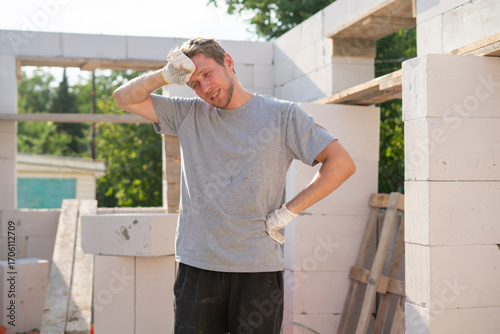 A construction worker in a gray shirt wipes sweat from his forehead while surrounded by concrete blocks on a hot day at the construction site, visibly exhausted from the labor.