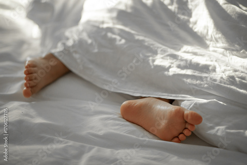 Kid bare feet covered in a white blanket. Child lies on the bed in the bedroom at home