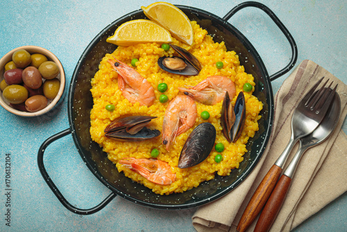 Seafood paella with rice, shrimp, mussels, peas, and lemon wedges served in a black traditional pan on a dark rustic background with olives and cutlery, Mediterranean meal