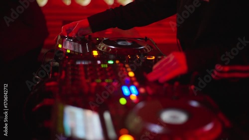 DJ hands manipulating a turntable mixer in a nightclub, colorful lights and red lights, blurred background, techno and electronic music atmosphere, buttons and knobs in focus, night party vibe.