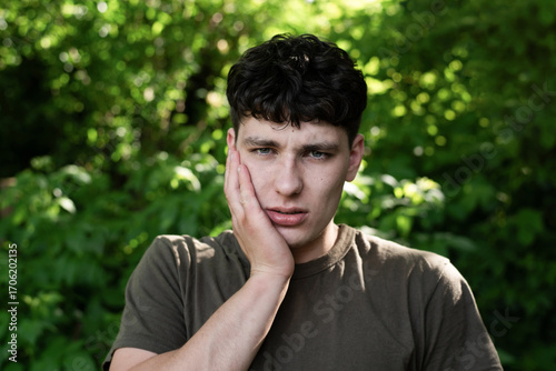 Worried young man holding face, his tooth hurts badly, he needs to go to dentist. A young man appears thoughtful and concerned while outdoors, expressing stress or pain