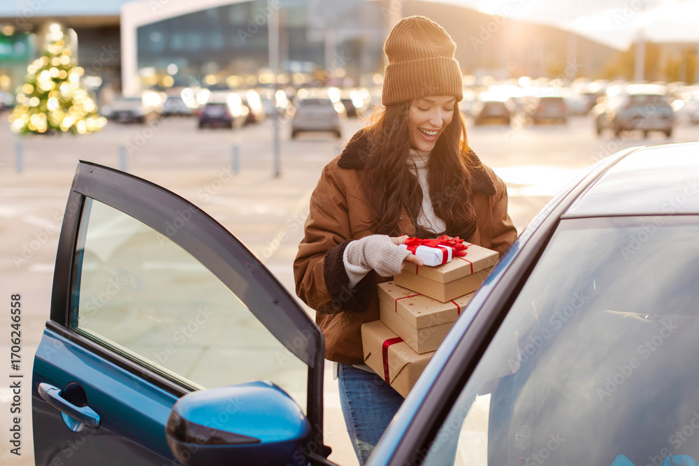 Naklejka premium Excited woman putting Xmas gift boxes into car after shopping in mall, enjoying holidays preparation, standing at outdoor parking lot