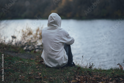 Lonely young man sits on the river bank in cloudy autumn weather, back view