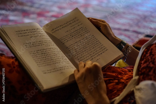 Reading Arabic poetry book on colorful traditional rug outdoors, blurred mountain landscape background, Nizwa, Ad Dakhiliyah Region, Oman