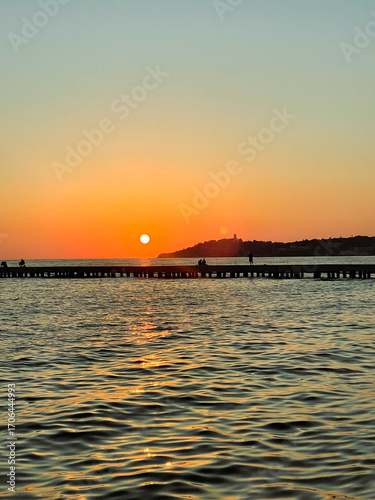 people enjoying sunset at the beach golden sunset illuminates the coastal sea waves and rugged rocks in Kusadasi, Turkey, creating a warm and serene atmosphere against the backdrop of the Aegean Sea
