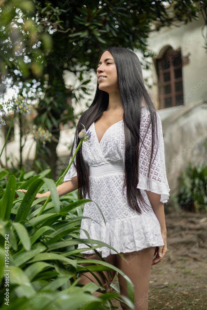 Naklejka premium Elegant young woman with long black hair wearing a white lace dress, standing outdoors while smelling a plant in a rustic colonial garden.