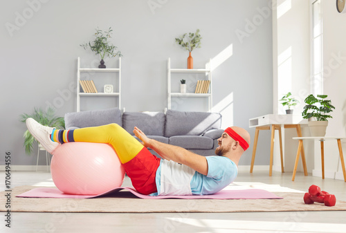 Fototapeta Naklejka Na Ścianę i Meble -  Funny young man in sportswear having home workout lying on floor on yoga mat with fit ball. Sporty male person exercising at home. Sport, fitness, health and home training concept.