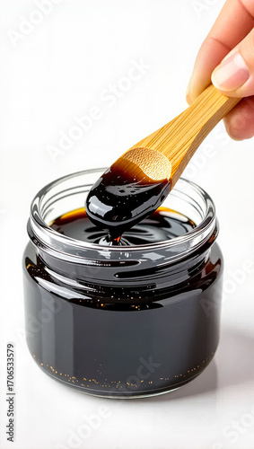 Hand holding wooden spatula, scooping shilajit resin from a glass jar, bright clean background, health and wellness concept.