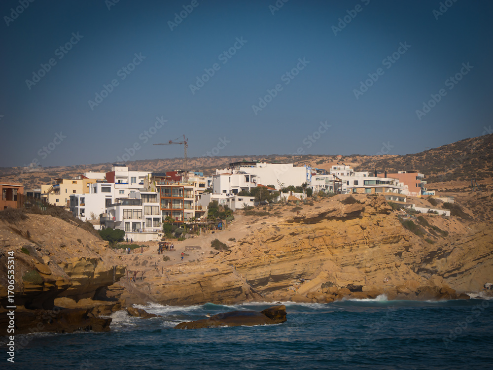 Fototapeta premium Village with modern, white buildings built along rocky, sandy cliffside. Scenic coastal view of holiday destination of Imsouane, Morocco where gentle waves of Atlantic Ocean roll onto the shore below