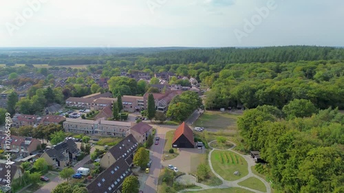Aerial view of a suburban area bordering a forest, featuring residential buildings, a red-roofed structure, parking, and community gardens. The layout blends housing, agriculture, and nature.