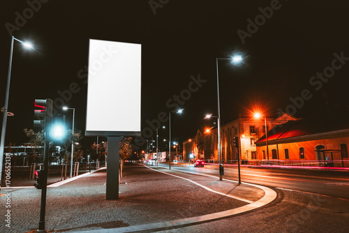 Large blank vertical billboard mockup illuminated at night on urban street with glowing streetlights, traffic lights, moving car lights and historic buildings in the background