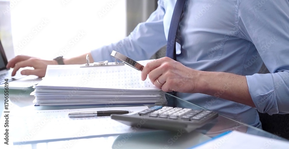Fototapeta premium A businessman is using a calculator and a magnifying glass to analyze financial documents at his glass desk in a modern and fair office setting, focusing on aspects of audit and taxes in business