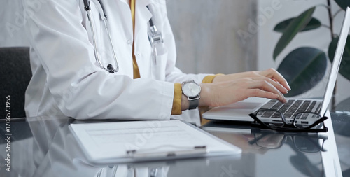 Fotografija Doctor woman wearing white medical coat over a yellow blouse is working on laptop with stethoscope on the glass desk
