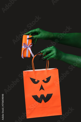 Green hands of witch with gift box and shopping bag on black background. Hall...