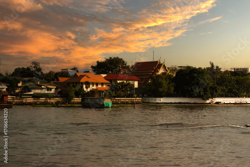 Traditional riverside Chao Phraya river surrounded by homes and high temple on golden details in Bangkok, Thailand at sunset