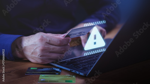 Customer holding a credit card while using a laptop computer with warning symbol, representing online payment risk, financial fraud, online shopping and identity theft during digital transactions.