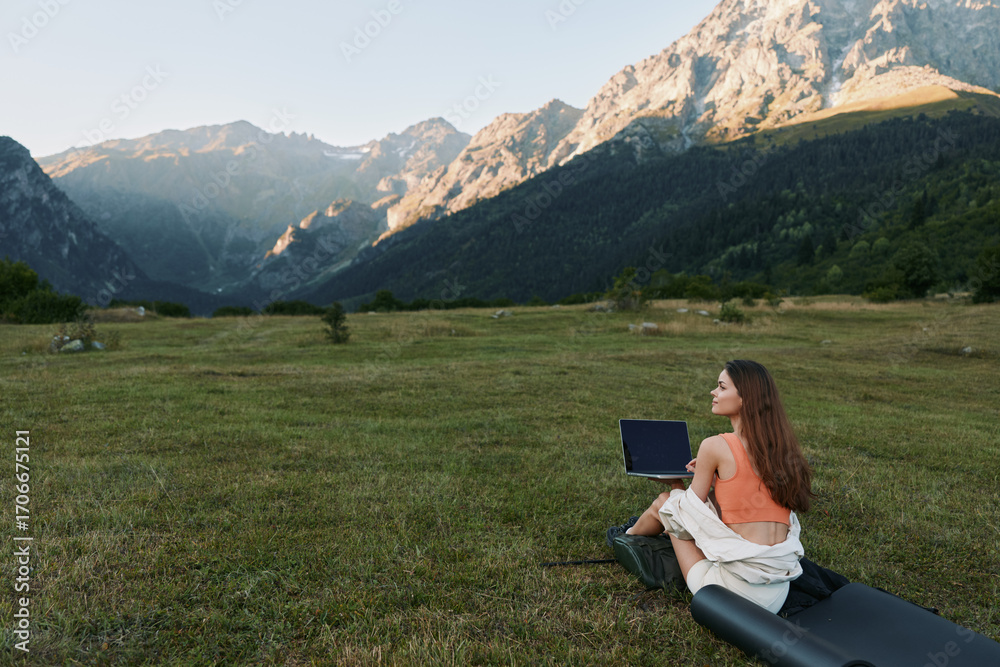 Fototapeta premium A woman sits on a grassy meadow with a laptop in a remote mountain setting. She appears focused and calm, enjoying a peaceful, productive outdoor workspace with expansive scenery and blue sky above.
