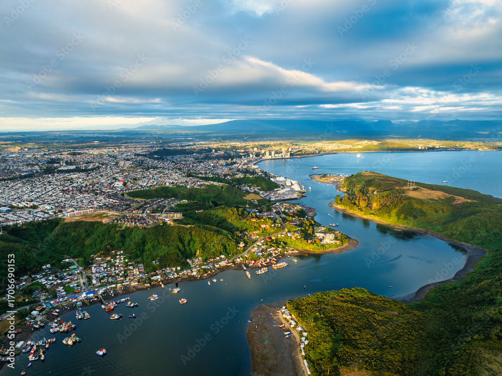 Fototapeta premium An aerial view of Puerto Montt Canal de Tenglo during the afternoon, with a mix of golden light and dramatic shadows on the landscape under a cloudy sky.