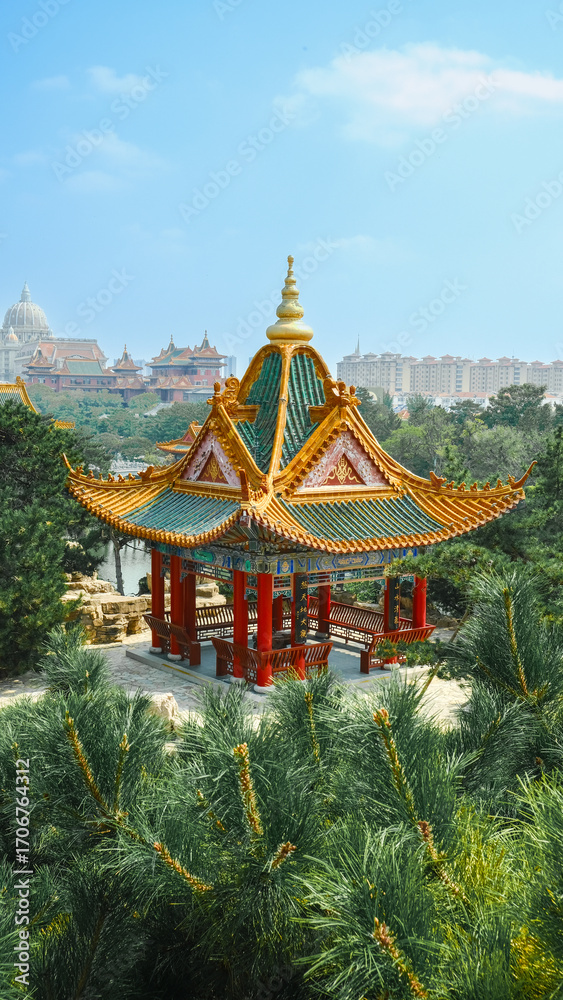 Naklejka premium Traditional Chinese temple in Penglai, Shandong, with red pillars, golden roofs, and ornate details, surrounded by lush greenery and a serene natural setting.