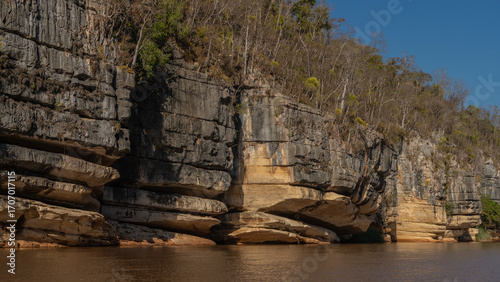 Coastal eroded limestone cliffs. There is green vegetation on the steep slopes. The blue sky. A calm red-brown African river. Madagascar. Manambolo river.