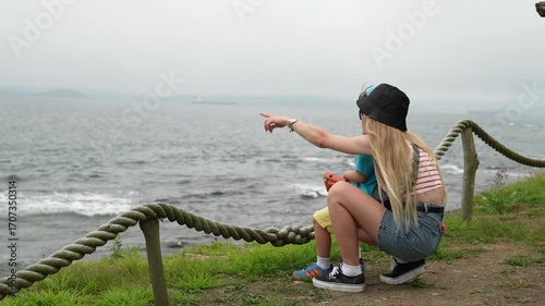 A mother and son are sitting by a rope bridge. The mother is showing her child something in the distance.