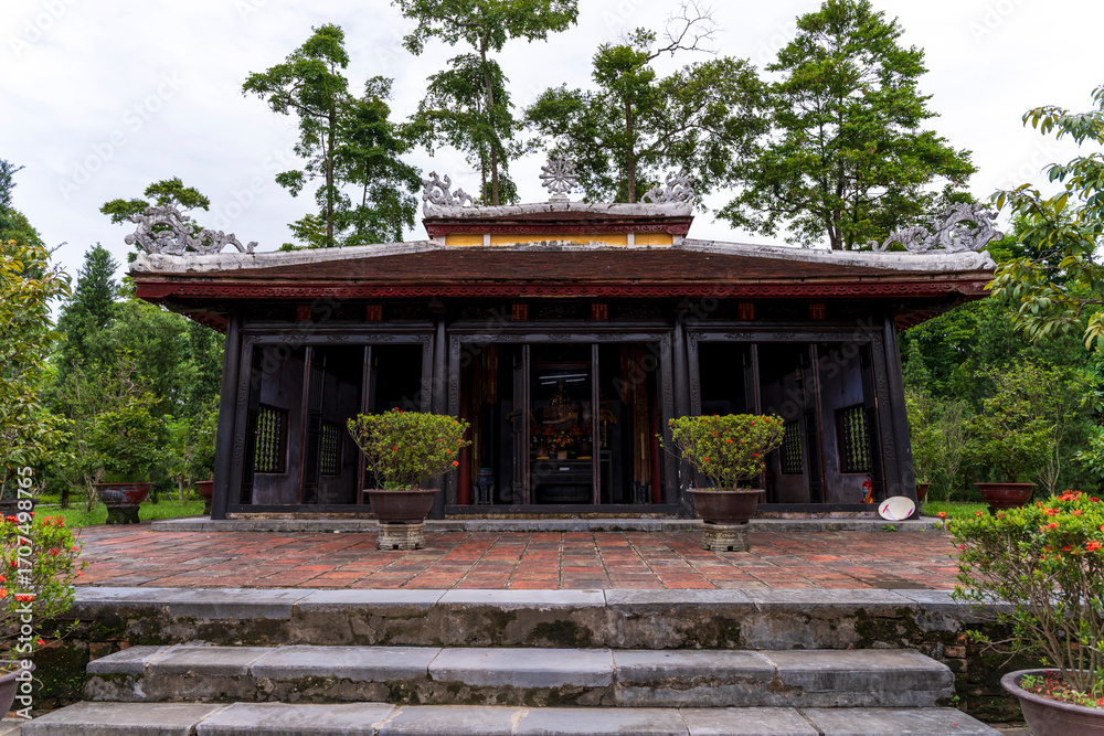 Obraz premium A brick pathway leads symmetrically toward a traditional temple building, flanked by manicured cypress trees and green lawns, creating a peaceful and spiritual atmosphere.