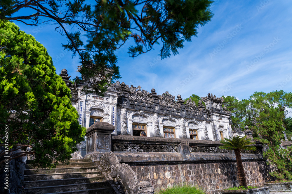 Fototapeta premium A broad view of the Khai Dinh Tomb's intricate, ornate exterior, with its detailed stone carvings and grand staircase under a striking blue sky.