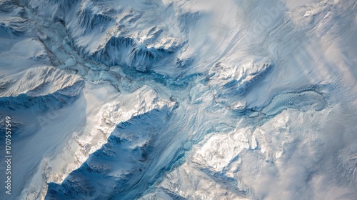Aerial view of a snow covered mountain range with glaciers and rivers in winter season with a blue hue and sunlight
