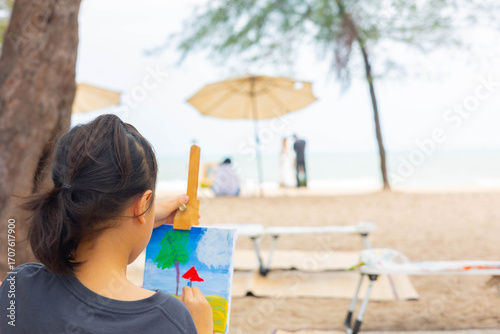 Over-the-shoulder view with blurred background of a young girl painting a seascape on an easel at beautiful beach, representing childhood creativity, outdoor learning, and a peaceful artist hobby.