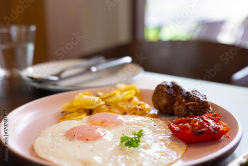 Selective focus close-up of breakfast plate with fried eggs, potato wedges, grilled tomato, and meatballs in morning. Food photography, restaurant menu design, healthy brunch concepts.