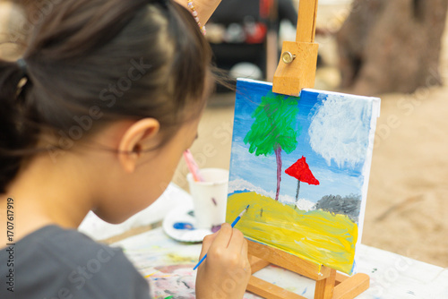 Close-up and selective focus of young girl painting a palm tree and sea with beautiful color on canvas. This shows creativity of a child's artistic hobby in a natural, outdoor beach setting.
