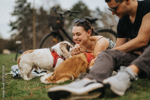 Photography A cheerful couple sitting on grass in a park interacting with their dogs on a pleasant day, surrounded by a casual outdoor atmosphere with bikes and natural scenery in the background