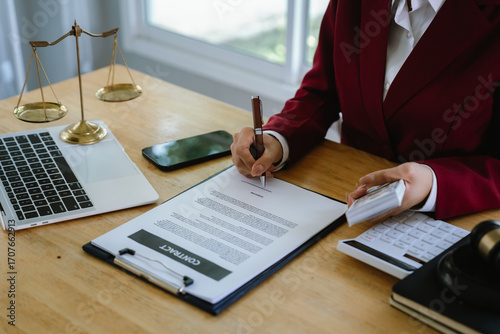 A professional reviews legal and financial documents, using a calculator, money stack, and laptop beside justice scales in office.