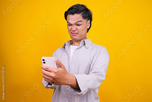Young Asian Man Reacting With Disgust To Something On Smartphone Isolated On Yellow Background