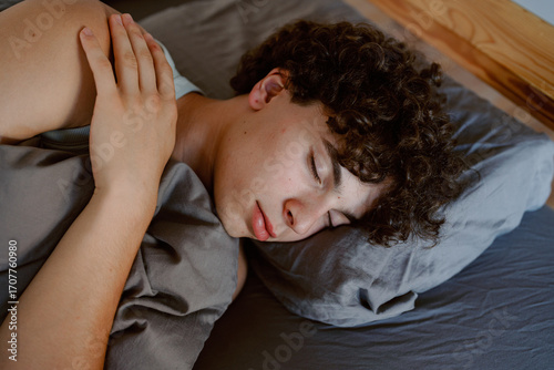 A young man is lying on his side in a softly lit bedroom, sleeping soundly on a comfortable bed. He has curly hair and looks relaxed under a cozy blanket.