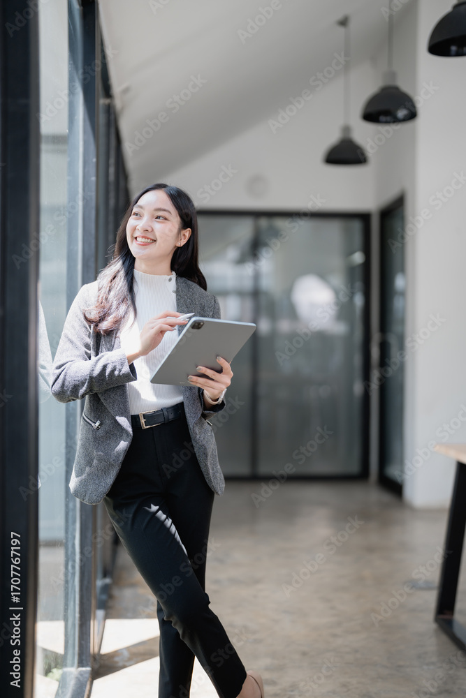 Fototapeta premium Young asian businesswoman using digital tablet and smiling while leaning on a large window in a modern office, she is wearing a gray jacket and black pants