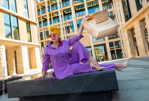 woman with craft shopping bags in the city. The concept of fashionable Consumerism and ecology. An adult Asian woman in a purple suit with red and yellow accents enjoys shopping . a sunny day.