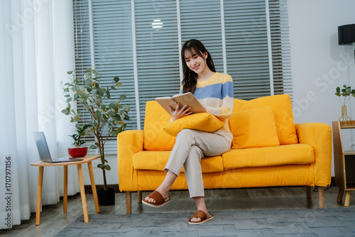 Beautiful young asian woman reading a book and smiling while sitting on a comfortable sofa in her modern living room, enjoying a relaxing afternoon at home