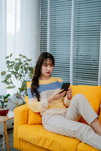 Happy young asian woman relax on comfortable couch at home texting or chatting messaging on the phone, smiling girl use cellphone, online message, shopping online from home