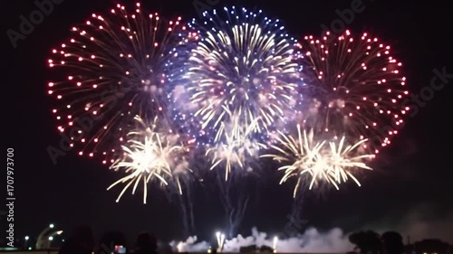 Bursting fireworks illuminate the night sky with vibrant colors and intricate patterns, captured from a distance with onlookers below