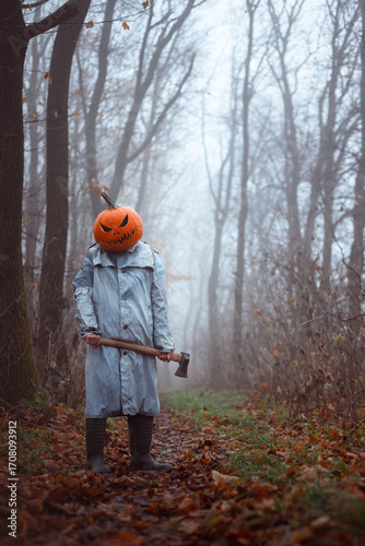 Haunting scarecrow with axe and carved pumpkin head stands still in misty autumn forest. Tattered cloak and fog add to the eerie Halloween scene and horror vibe