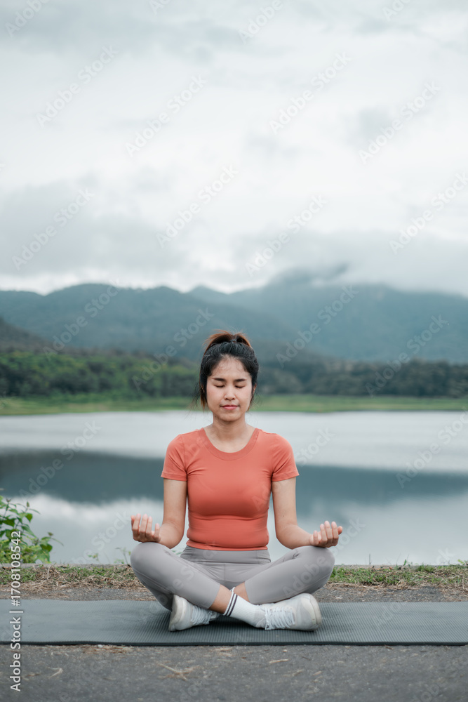 Fototapeta premium A woman practices meditation by a tranquil lake with mountains in the background, creating a peaceful and serene atmosphere.