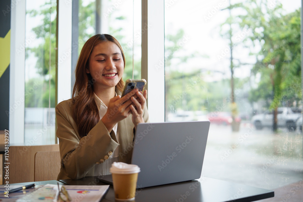 Obraz premium Young female entrepreneur checking mobile messages during remote work session at cafe with laptop and charts.