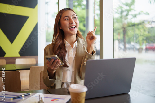 Excited businesswoman holding smartphone and pointing upward with a smile while working on laptop in a cafe.