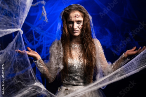 Obraz na plátne Unhappy creepy woman with horrible scary Halloween zombie makeup in white dress says why do you want, standing on blue background surrounded spiderweb