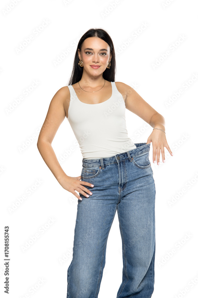 Obraz premium Studio shot of young woman with long dark hair wearing white tank top and loose blue jeans holding waistband to show fit on white background.