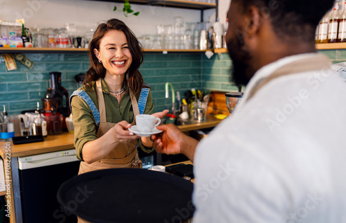 Female barista working in a coffee shop at the counter and gives a coffee to a male waiter to serve to the customer.