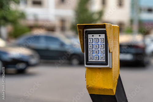 Electronic keypad device mounted on yellow post in outdoor parking area, secure access control technology with blurred cars in background.